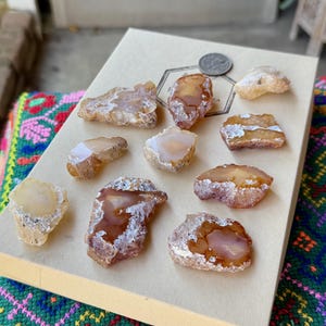 Può includere: Una collezione di pietre di agata lucidata nei toni del marrone, beige e bianco, disposte su una scatola beige. Una moneta d'argento è posta sulla scatola. Le pietre variano per dimensioni e forma, mostrando motivi e texture naturali. Lo sfondo è un tessuto colorato.