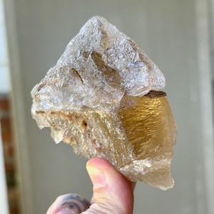 May include: A raw, natural crystal formation with a translucent, light brown hue. The crystal has a rough, uneven texture and a golden-yellow section. The overall shape is irregular, with sharp edges and a crystalline structure.