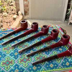 May include: Six smoking pipes arranged in a diagonal row. Each pipe features a smooth, deep red wooden bowl and a long, dark brown, textured stem. The pipes are positioned on a vibrant, multi-colored fabric surface with a geometric pattern.