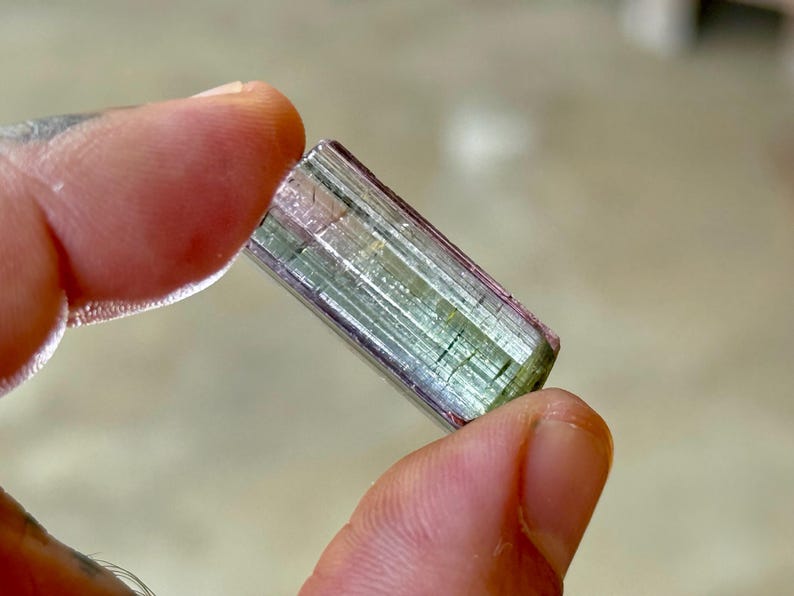 May include: A multi-colored tourmaline crystal, held between fingers. The crystal is a rectangular prism with visible layers of green, pink, and clear sections. The gemstone is approximately 1 inch long.