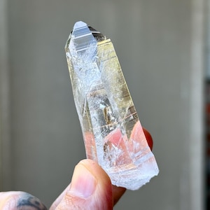 May include: A clear quartz crystal with a pointed tip and a rough, textured base. The crystal is held up against a blurred background. The quartz has internal lines and inclusions, and it appears to be translucent.