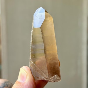 May include: A translucent, light brown smoky quartz crystal point with a hexagonal shape and a pointed termination. The crystal features visible horizontal growth lines along its smooth, elongated faces and a clear, glassy texture throughout.