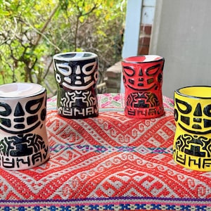 May include: Four colorful ceramic tiki mugs with a tribal design. The mugs are in shades of beige, black, red, and yellow, each with a unique face and body pattern. They are arranged on a red and white patterned textile.