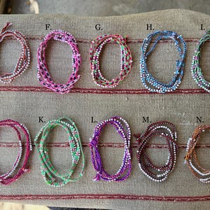 May include: An assortment of colorful beaded bracelets in various shades, including pink, blue, green, and purple. Each bracelet is intricately designed with small beads and woven threads, displayed on a textured surface.