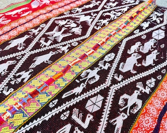Op de afbeelding: Close-up van een kleurrijk geweven textiel met geometrische en dierlijke patronen. Het textiel heeft een donkerbruine achtergrond met witte vogel- en mensachtige figuren. Felle kleuren zijn rood, geel en oranje. Waarschijnlijk een deken of wandkleed.
