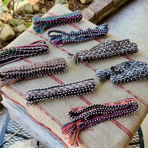 May include: A collection of colorful woven belts with intricate patterns and beaded details. The belts feature a variety of colors, including red, purple, blue, and brown. Each belt has a tassel at the end.