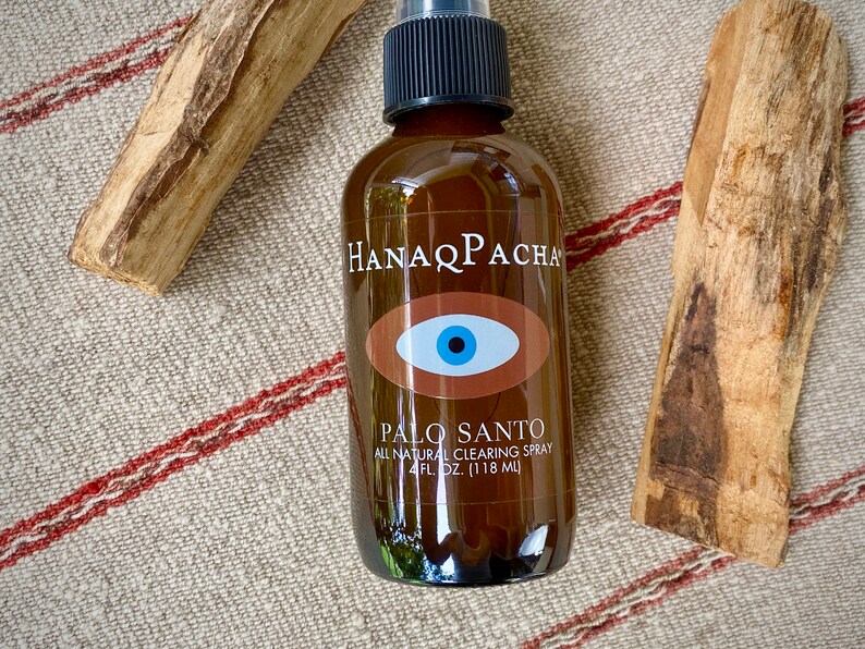 Peut inclure: Une bouteille en verre brun avec un vaporisateur noir et une &eacute;tiquette qui indique "Hanaq Pacha" et "Palo Santo All Natural Clearing Spray 4 fl. oz. (118 ml)". L'&eacute;tiquette pr&eacute;sente &eacute;galement un &oelig;il brun avec un iris bleu. La bouteille est pos&eacute;e sur un tissu ray&eacute; beige et rouge.