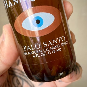 Peut inclure: Une bouteille en verre brun avec une &eacute;tiquette indiquant "PALO SANTO ALL NATURAL CLEARING SPRAY 4 FL. OZ. (118 ML)". L'&eacute;tiquette pr&eacute;sente &eacute;galement un grand &oelig;il bleu stylis&eacute; avec une pupille noire.
