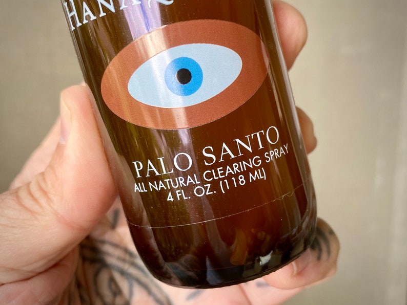 Peut inclure: Une bouteille en verre brun avec une &eacute;tiquette indiquant "PALO SANTO ALL NATURAL CLEARING SPRAY 4 FL. OZ. (118 ML)". L'&eacute;tiquette pr&eacute;sente &eacute;galement un grand &oelig;il bleu stylis&eacute; avec une pupille noire.