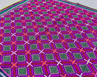 May include: A colorful square rug with a geometric pattern in shades of pink, green, red, white, and black. The rug is made of a woven fabric and has a fringed edge.