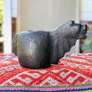 Puede incluir: Una escultura en forma de oso gris oscuro con una superficie texturizada. El oso está colocado sobre un textil estampado colorido con tonos rojos, blancos y azules. La escultura tiene un área ahuecada en su parte posterior.