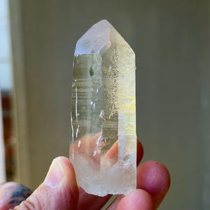 May include: A clear quartz crystal point, held in a hand. The crystal is transparent with a slightly cloudy appearance and a pointed tip. The crystal has visible internal structures and imperfections.