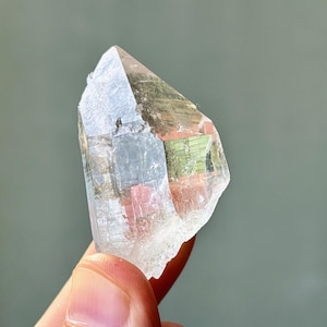 Op de afbeelding: Een heldere kwartskristal met een veelzijdige, puntige vorm. Het kristal heeft een doorschijnende kwaliteit met interne kleurvariaties van roze, groen en grijs. Het kristal wordt in een hand gehouden.