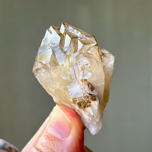 May include: A natural, raw quartz crystal cluster with a light tan colour. The crystal has multiple points and facets, with some areas appearing translucent. The overall shape is irregular, with a rough texture and some darker brown inclusions.