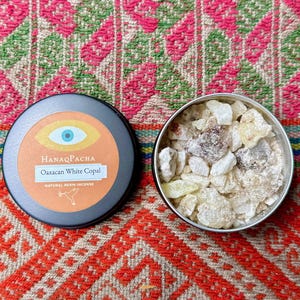 May include: A black tin with an orange label and the text "HanaqPacha Oaxaca White Copal Natural Resin Incense" sits next to a silver tin filled with white and tan resin pieces. The tins are on a colourful patterned textile.