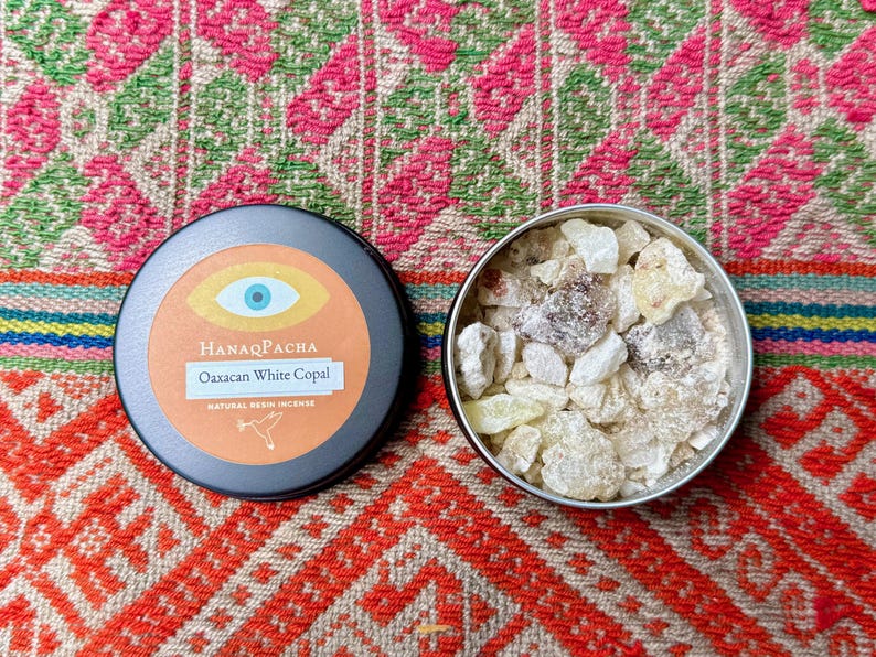 May include: A black tin with an orange label and the text "HanaqPacha Oaxaca White Copal Natural Resin Incense" sits next to a silver tin filled with white and tan resin pieces. The tins are on a colourful patterned textile.