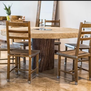 May include: A round wooden table with a natural wood finish and a rough-hewn top. The table is surrounded by six wooden chairs with slatted backs and metal accents. The chairs are arranged around the table in a circular pattern.