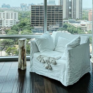 May include: A white linen armchair with a white throw blanket and a wooden stump side table. The chair is positioned in front of a large window with a view of a city skyline.