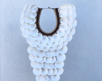 May include: A white shell necklace with brown beads hangs from a black stand. The necklace is made of many small, round shells arranged in a cascading pattern.