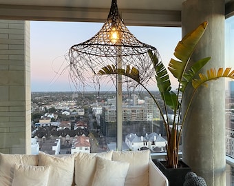 May include: A white couch with multiple pillows sits in front of a large window with a view of a city skyline. A woven pendant light hangs from the ceiling above the couch. A large potted plant with green leaves is to the right of the couch.