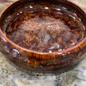 May include: A small, brown ceramic bowl with a dragonfly design. The bowl has a glossy finish and a speckled brown glaze.
