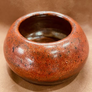 May include: A small, round, terracotta-colored ceramic bowl with a speckled glaze. The bowl has a slightly rounded lip and a smooth, glossy finish.