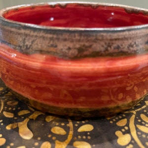 May include: A small, red ceramic bowl with a brown rim and a subtle pattern. The bowl is handmade and has a rustic, earthy feel.