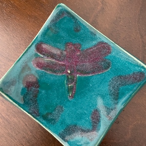 May include: A square teal ceramic dish with a raised dragonfly design in a deep purple color.