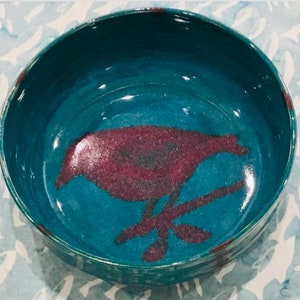May include: A handmade ceramic bowl with a teal blue interior and exterior. The bowl features a maroon bird design on the inside. The bird is perched on a branch. The bowl is round and has a glossy finish.