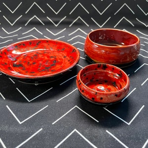 May include: Three red and black ceramic bowls with a speckled glaze. The bowls are nested inside each other, with the largest bowl on the bottom and the smallest bowl on top.