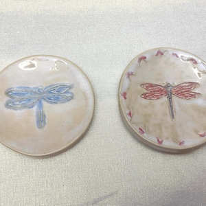 May include: Two round ceramic coasters. One coaster features a blue dragonfly design, the other a red dragonfly. Both coasters have a cream-colored background and are likely handmade. Decorative and functional.