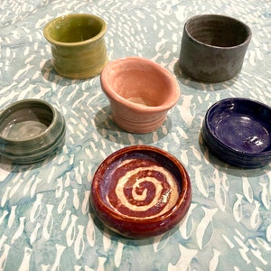 May include: Six small ceramic bowls in various colors and designs. The bowls are arranged on a light blue surface with a white pattern.