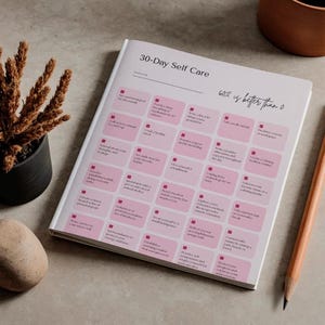 Op de afbeelding: Een 30-daags zelfzorgdagboek met roze vakjes en tekst. Een potlood ligt naast het open dagboek. Een kleine potplant en een decoratieve steen staan ook op de foto.