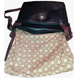 Puede incluir: Un bolso bandolera de cuero negro con costuras y bordes en rojo oscuro. El bolso presenta un emblema met&aacute;lico circular de tono plateado en la solapa frontal. El interior est&aacute; forrado con tela beige claro con un patr&oacute;n de c&iacute;rculos blancos.