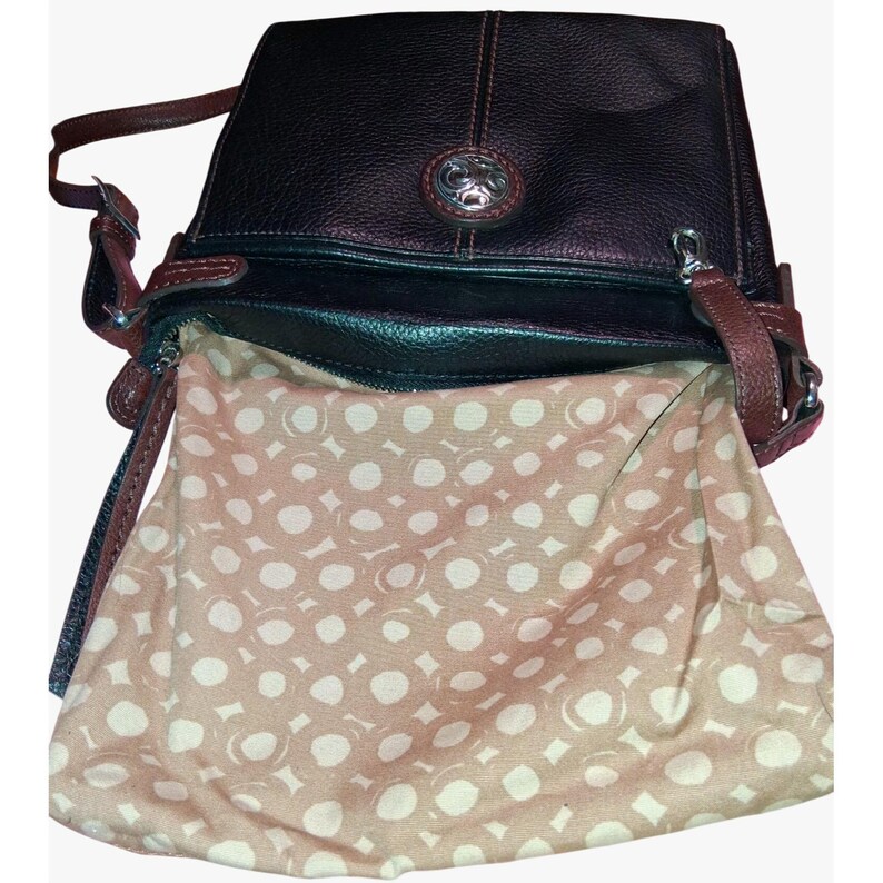 Puede incluir: Un bolso bandolera de cuero negro con costuras y bordes en rojo oscuro. El bolso presenta un emblema met&aacute;lico circular de tono plateado en la solapa frontal. El interior est&aacute; forrado con tela beige claro con un patr&oacute;n de c&iacute;rculos blancos.