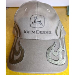 May include: A tan baseball cap featuring the John Deere logo and a leaping deer emblem embroidered in dark thread on the front panel. The cap includes decorative flame-like patterns stitched in a darker tan thread across the brim and sides.