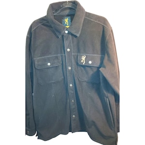 May include: A black button-up shirt with two chest pockets and a collar. The shirt features a small logo on the chest and a tag on the collar. The shirt is hanging on a hanger against a white background.