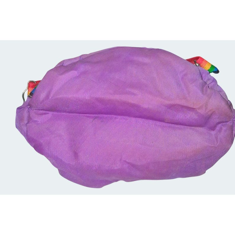May include: A lavender-colored shoulder bag with a rainbow strap. The bag is made of a soft, possibly nylon, material. The strap is a woven rainbow pattern. The bag is a rounded shape.