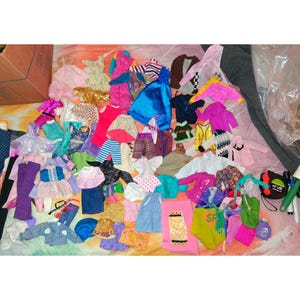 Op de afbeelding: Een verzameling poppenkleding in verschillende kleuren en stijlen. Het assortiment omvat jurken, jassen, rokken, broeken en tops. Kleuren variëren van roze, blauw en geel tot groen, paars en bruin. Sommige items hebben patronen.