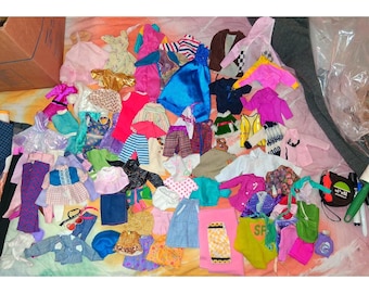 Può includere: Una collezione di vestiti per bambole in vari colori e stili. L'assortimento comprende abiti, giacche, gonne, pantaloni e top. I colori vanno dal rosa, blu e giallo al verde, viola e marrone. Alcuni articoli hanno motivi.