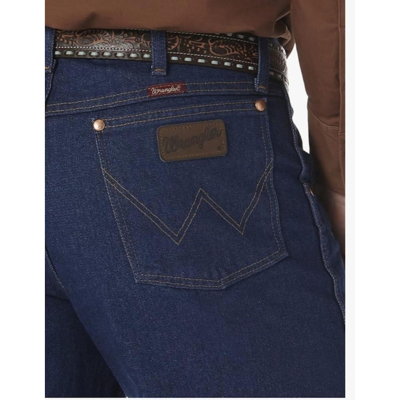 May include: Dark blue denim jeans with the Wrangler logo on the back pocket. The jeans have copper-colored rivets and a brown leather patch. A brown leather belt with a decorative pattern is visible.