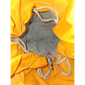 May include: A bright yellow drawstring bag with a light gray interior. The bag's opening is visible, revealing the inside. Beige rope is used for the drawstring closure, and metal grommets reinforce the holes. The bag appears to be made of a durable fabric.