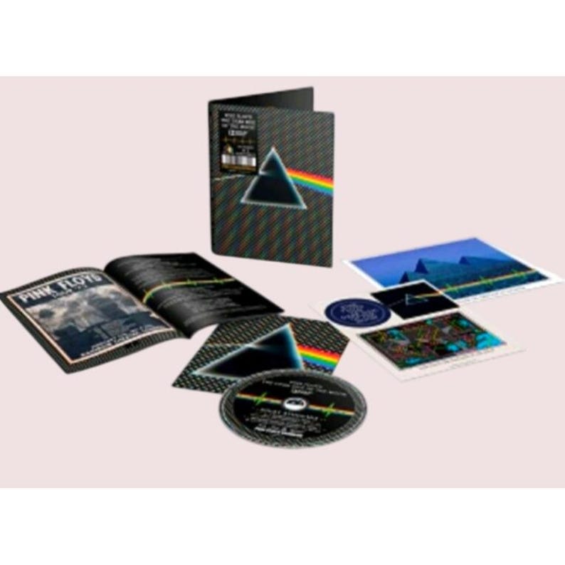 May include: A Pink Floyd The Dark Side of the Moon box set featuring a black textured case with a prism graphic, an open booklet, a circular disc, and several square art prints. The prism design displays a white light beam refracting into a rainbow.