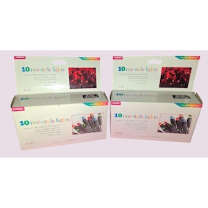 May include: Two boxes of NOMA 10 rice-style lights. Each box has a white background with the product name in teal and a picture of the lights. The lights are multi-colored and are designed for wreaths, garlands, plants, and miniature trees.