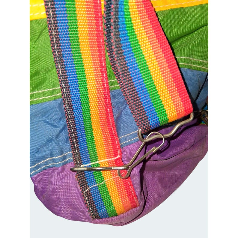 May include: Close-up of a colorful bag with a rainbow strap. The strap is attached to the bag with a metal clip. The bag is made of various colors, including green, blue, purple, and yellow.