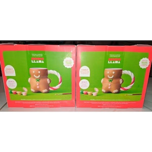 May include: Two boxes featuring a gingerbread man-shaped mug with a candy cane handle. The mug is brown with white icing details and a green bow. The boxes are red and green with the text "LLAMA".