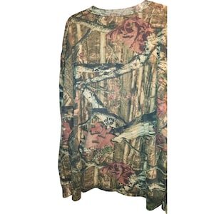 May include: Long-sleeved camouflage shirt in shades of brown, green, and tan. The shirt features a crew neck and a pattern of branches and leaves, typical of hunting apparel.