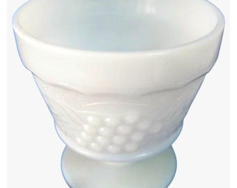 May include: A white milk glass footed bowl featuring a raised, embossed pattern of grape clusters and leaves around the exterior. The bowl has a wide, circular opening, a smooth rim, and a sturdy pedestal base, showcasing a semi-translucent finish.