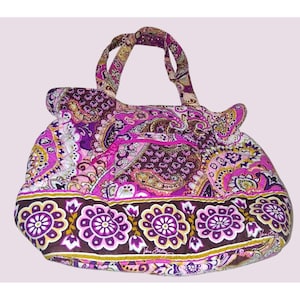 Puede incluir: Un bolso tote acolchado con un estampado de cachemir y floral en tonos rosa, morado y marrón. El bolso tiene dos asas y un bolsillo con cremallera. La parte inferior del bolso presenta una fila de grandes flores moradas y blancas.