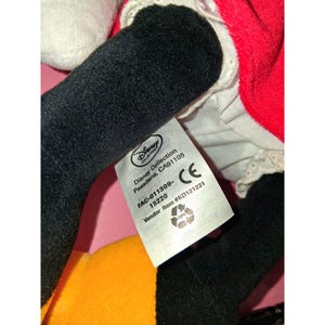 May include: A plush toy featuring black arms and a portion of a red dress. A tag from the Disney Collection is visible, with text including "Pasadena, CA91105" and "FAC-011309-". The toy has a white and yellow section.